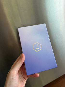 A hand holds a light purple/lavender rectangular box featuring a minimalist gold line-art logo of a cat or fox face, photographed against a gray fabric background.