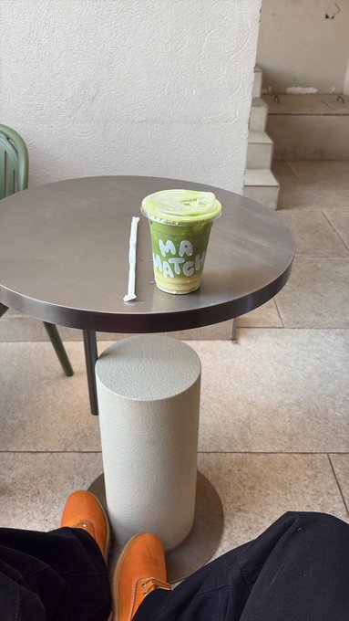 A matcha latte in a green cup labeled 'MATCHA' sits on a round metal table with a straw, while a person in orange slippers sits nearby.