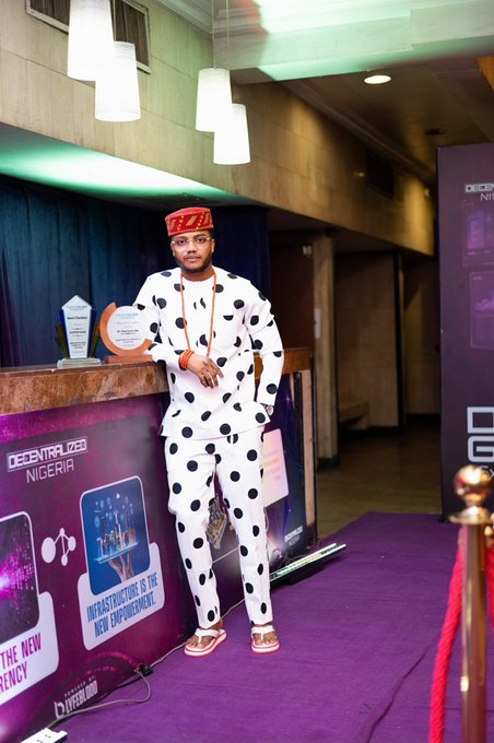 A man in a white polka-dot traditional outfit and red cap poses next to a "Decentralized Nigeria" branded booth featuring text like "Infrastructure is the New Empowerment" and "The New Currency," as he displays an award he received for Web3 Marketing Personality of the Year from the event organizers.