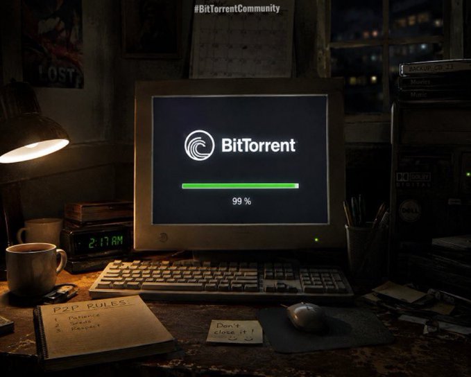 A dimly lit desk setup features an old CRT monitor displaying the BitTorrent logo with a nearly complete green progress bar at "99%," while a notebook labeled "P2P RULES!" sits beside a coffee mug. The scene visually echoes the post's message by symbolizing BitTorrent's ongoing evolution from its file-sharing roots toward its new BTTC blockchain and Web3 infrastructure.