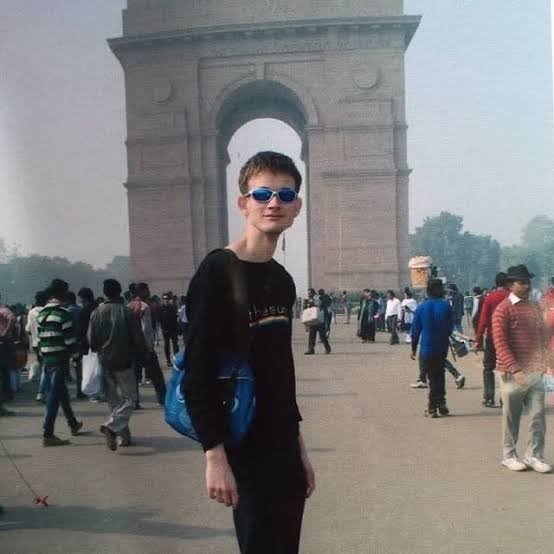 A young boy with a backpack and blue sunglasses stands in front of India Gate in a hazy, crowded plaza.