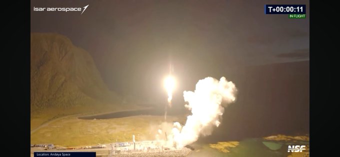 A nighttime rocket launch from Andøya Space by ISAR Aerospace, with the bright engine flame illuminating the launch pad and surrounding terrain, marked 'IN FLIGHT' at T+00:00:11.