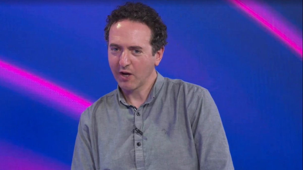 The image shows a man wearing a gray shirt, standing against a blue background with pink lines. The man appears to be speaking or giving a presentation, possibly at an event like a conference or a live stream. The caption of the image reads "This clip is poignant for current X conversations. As @Vet_X0 said yesterday, 'the more assets brought to the ledger, the better.' Focus on what is in the image and how it relates to the post".
