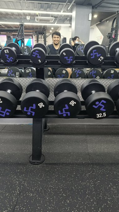 A man in a gym smiles from behind a large dumbbell rack filled with various weights, with the closest dumbbells marked 30 kg and 32.5 kg.