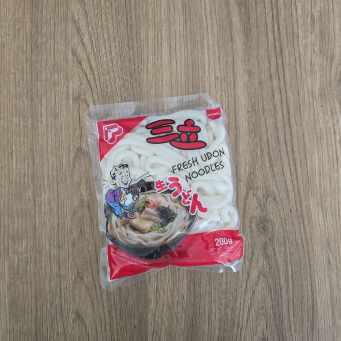 A 200g package of 'FRESH UDON NOODLES' featuring Japanese characters and a cartoon chef mascot, resting on a wooden surface.