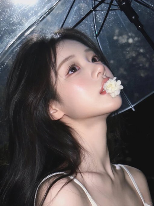 A young woman with long dark hair posing under a clear umbrella at night with a white flower near her lips.