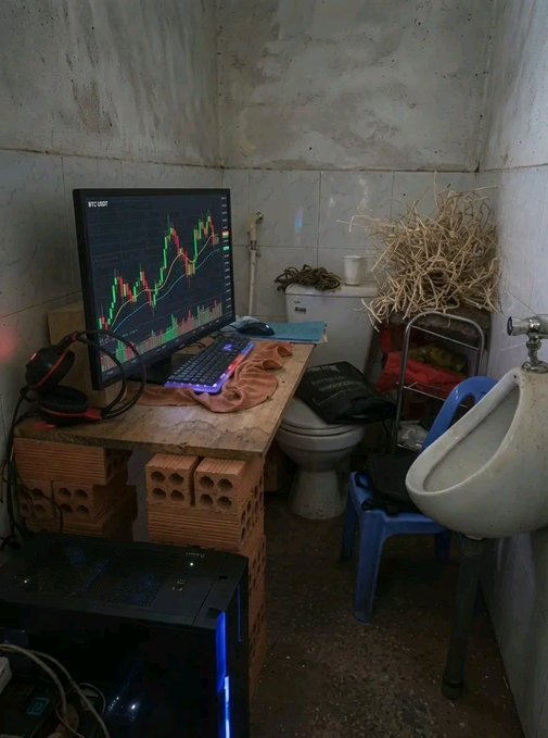 A humorous scene of a trading setup in a bathroom, with a computer monitor displaying a green and red candlestick chart on a makeshift desk of bricks, situated next to a toilet and urinal.