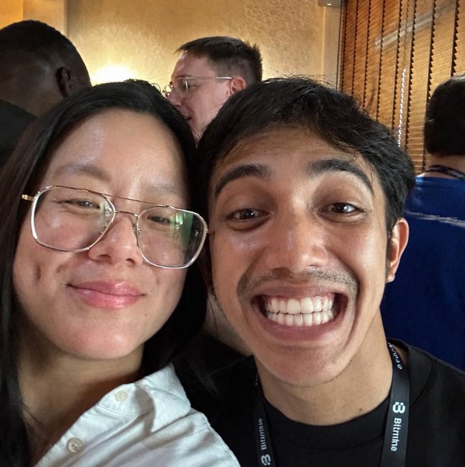 A selfie of two people smiling at the camera at what appears to be a conference; the woman on the left wears glasses and a white shirt, while the young man on the right wears a lanyard with 'Bitfinex' branding.