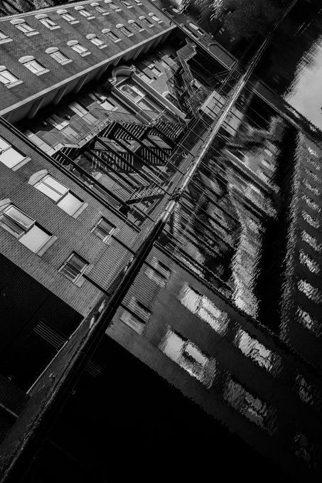 A dramatic black-and-white, tilted-angle photograph of urban apartment buildings featuring fire escapes, with strong reflections creating an abstract mirror effect across the architecture.