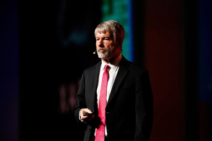 The image shows a man wearing a suit, speaking into a microphone at an event or conference. The man appears to be giving a presentation or speech, possibly discussing Bitcoin sales as part of Strategy's digital lending business model.
