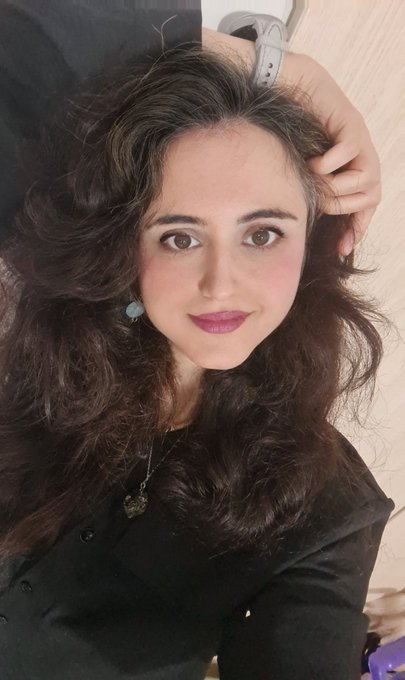 A close-up selfie of a young woman with long dark wavy hair, wearing a black shirt and a silver necklace, with her hand resting near her head.
