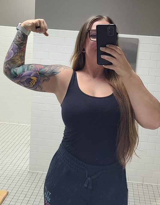 A selfie of a woman in a black tank top flexing her arm to display a colorful floral tattoo sleeve, taken in a tiled restroom.