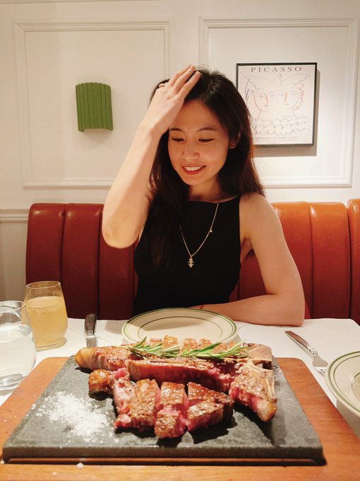 A woman sitting at a restaurant booth with a large sliced steak presented on a black stone platter in front of her, with a Picasso line art print on the wall behind her.