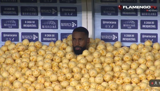 A man is humorously buried up to his neck in a pile of bread rolls ("pão de queijo") during what appears to be a Flamengo TV live broadcast, with a backdrop reading "MARACANÃ Tour" and "A CASA DO INESQUECÍVEL," tying into the cheesy, bread-filled post caption.