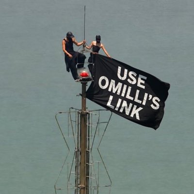 Two people in black clothing sit atop a tall structure, unfurling a large black flag that reads "USE OMILLI'S LINK" to promote the link shared in the post. The image visually reinforces the call-to-action, emphasizing the urgency and visibility of the message to help "Omilli" achieve his goal.