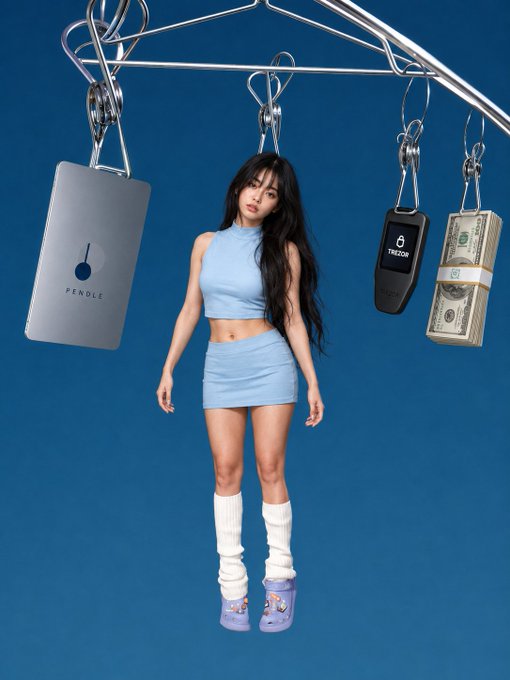 A young woman in a light blue crop top and skirt stands against a blue background with white leg warmers, surrounded by hanging objects including a "PENDLE" card and a "TREZOR" device.