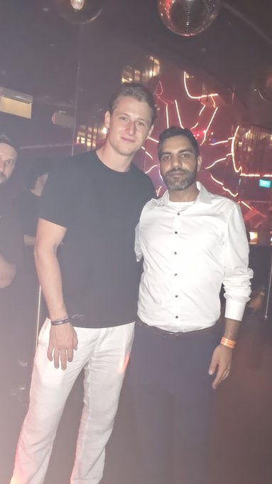 Two men pose together for a photo at what appears to be a nightclub or party venue with disco balls and neon lighting in the background.
