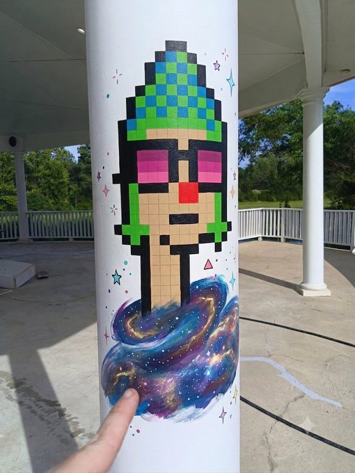 A column painted with a pixel-art style mural depicting a green-hatted figure with sunglasses emerging from a cosmic galaxy swirl, located in an outdoor pavilion.