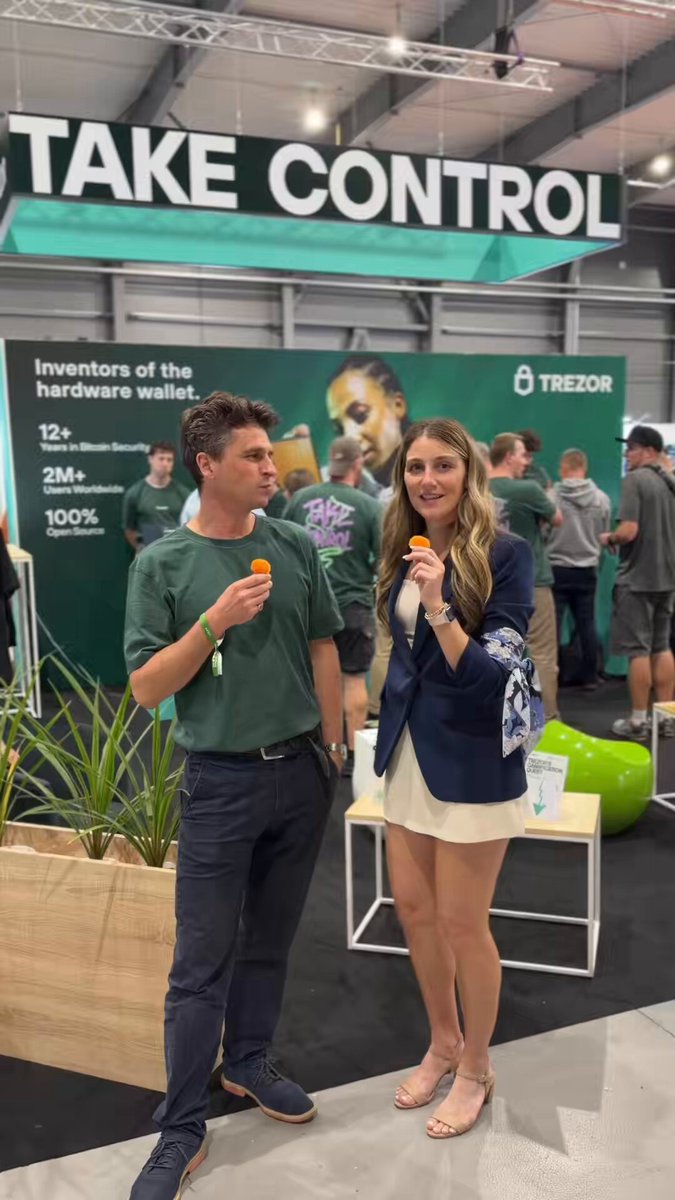 The image features a man and woman standing next to each other at an event with a green booth displaying information about Monero cryptocurrency. The woman has her arm around the man's waist as they pose for a photo, both holding orange lollipops in their hands. They are surrounded by several other people attending the same event or passing by on the street outside of the venue.