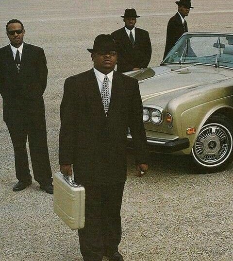 Four Black men in black suits, ties, sunglasses, and fedoras stand around a silver Rolls-Royce, with one holding a metallic briefcase in the foreground.