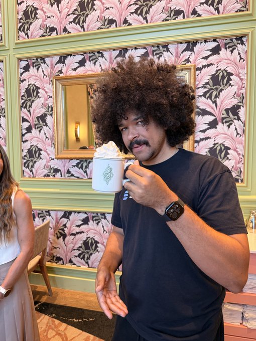 A man with a large afro and mustache drinks from a white mug with green text, wearing a dark t-shirt and black smartwatch, against a pink and black leaf-patterned wall with a mirror.