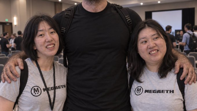 Two Asian women wearing matching gray 'MEGAETH' t-shirts posing on either side of a man in a black t-shirt at what appears to be a conference event.