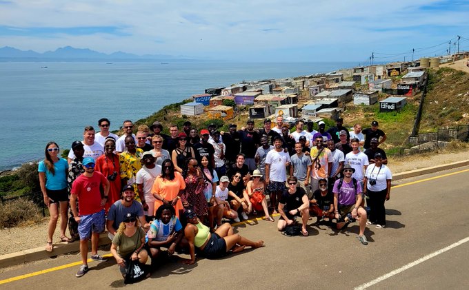 A large diverse group of people poses for a photo on a coastal road, with makeshift shacks and the sea visible in the background.