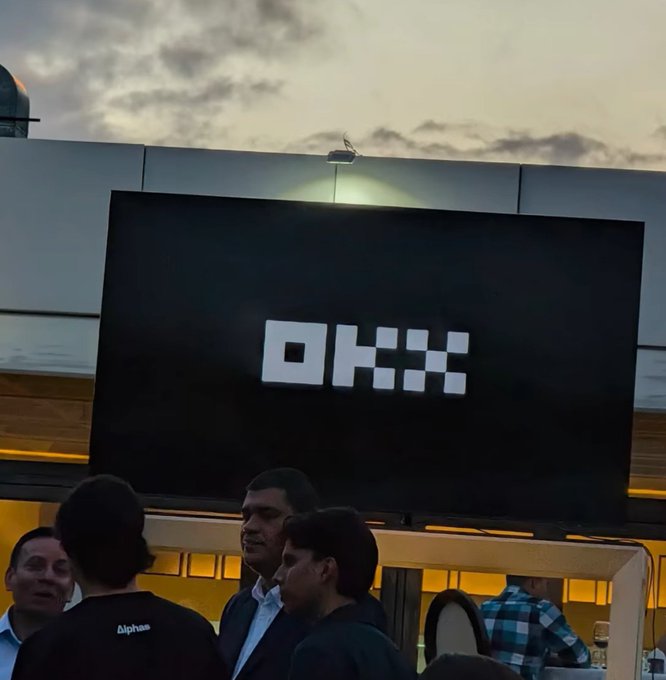 A nighttime scene at what appears to be an OKX-branded venue with a large illuminated OKX sign above people gathered at an outdoor event.