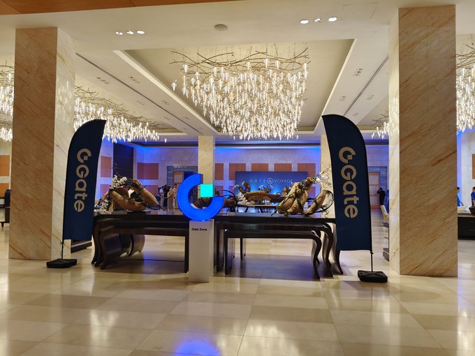 A luxurious hotel lobby features grand crystal chandeliers, marble pillars, and a central display with "Gate.io" banners and a blue logo, promoting a "GATE.IO VOYAGE" event.