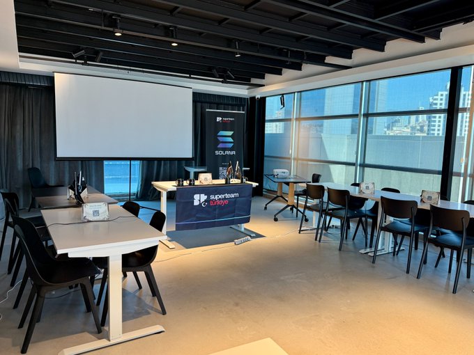 A meeting or event room features tables arranged with chairs, a projector screen, and a banner for 'SUPERTEAM SOLANA' with the Infinos logo, set against large windows with a city view.