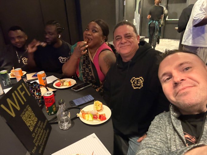 A selfie of a group of people seated at a table with food, drinks including Coca-Cola and Fanta, and a 'WIFI' QR code sign, appearing to be at a casual gathering or restaurant.