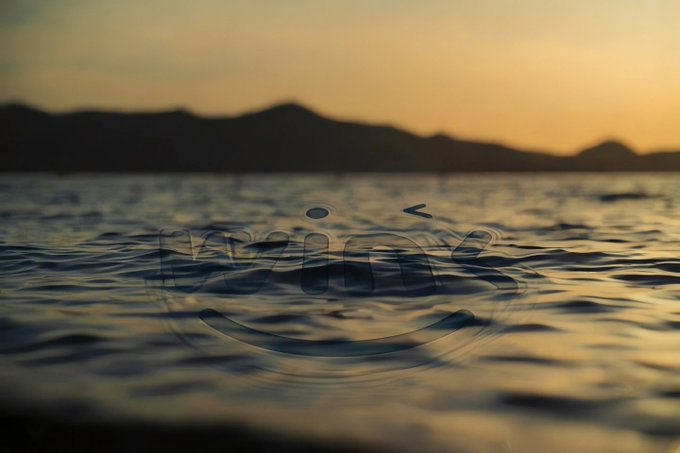 A serene view of rippling water at sunset with silhouetted mountains in the background and a faint smiley face watermark in the foreground.