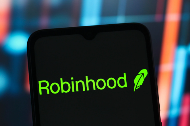 A close-up shot of a smartphone displaying the green 'Robinhood' logo and feather icon on its screen, with a blurred background of red and blue financial market charts.