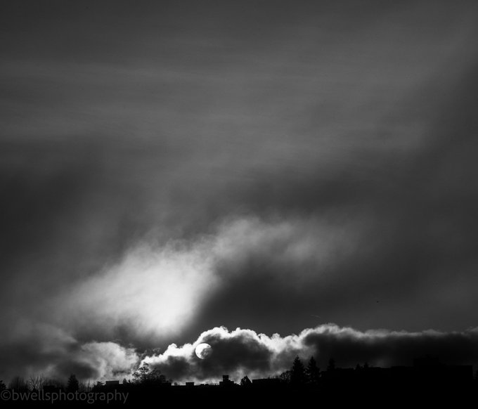 A black and white photograph by b-wells photography showing the moon partially hidden behind a dramatic, cloudy sky over a silhouetted treeline.