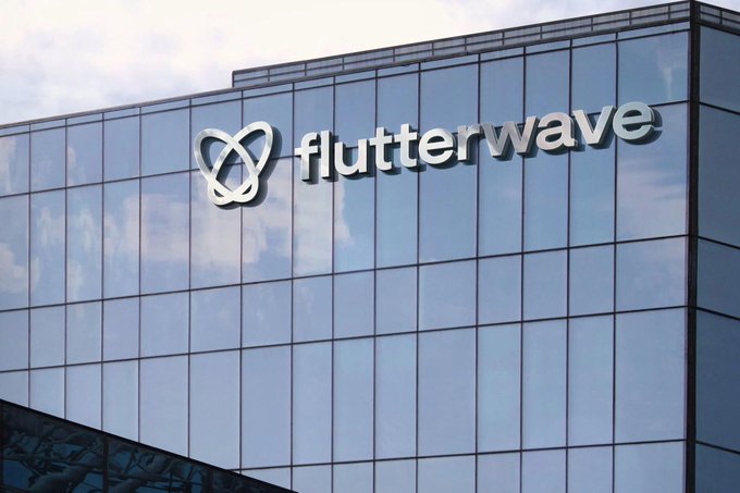 The image features a large glass building with a logo prominently displayed on its side, which reads "Flutterwave" in white letters. The building appears to be made of glass or have a reflective surface that enhances the visibility of the logo. The surrounding area seems to be an urban setting, as there are other buildings visible in the background.