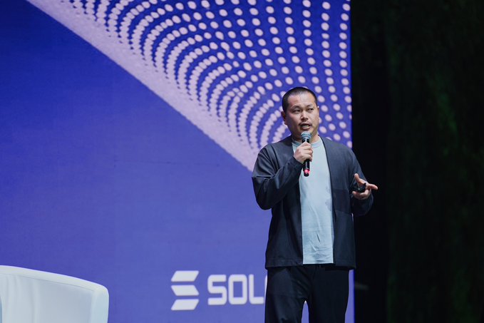 The image features a man standing at a podium, speaking into a microphone during an event. He appears to be delivering a speech or presentation, possibly related to the Solana Summit Kazakhstan. The backdrop of the scene includes a blue wall with white polka dots, and there are chairs placed around the area as well.
