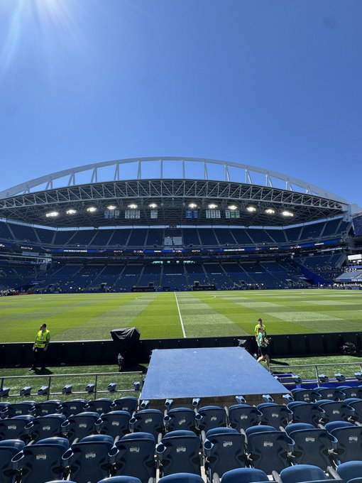 View from the stands of a large open-air stadium with empty blue seats, a green field, and bright sunlight streaming down.