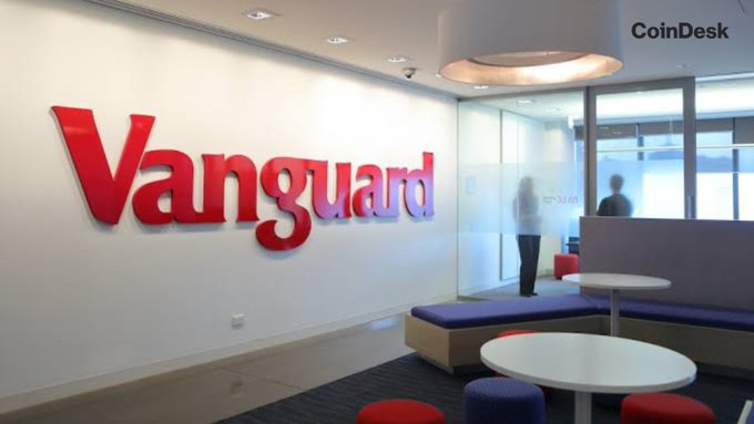 The image shows the interior of a Vanguard office, featuring a large red "Vanguard" logo on a white wall, with a modern reception area containing white tables, purple and red stools, and glass-walled offices. The "CoinDesk" watermark appears in the upper right corner. The image visually represents Vanguard's corporate identity, contextualizing the announcement of the asset manager's new Head of Digital Assets role as a notable strategic shift.