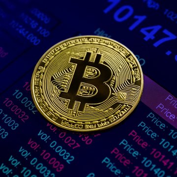 A gold physical Bitcoin token rests on a digital display showing live cryptocurrency market data, with visible numbers like "10147" and volume figures reflecting Bitcoin's active trading. The image visually reinforces the post's message about Bitcoin surging toward $87K amid intense FOMO-driven market activity and a major short squeeze.