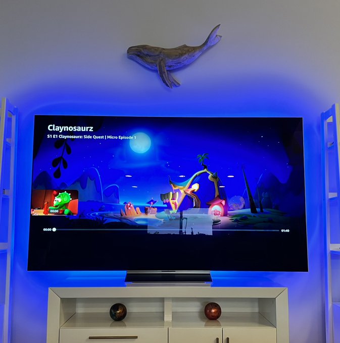A living room setup featuring a wall-mounted TV playing a video titled 'Claynosaurz: S1 E1 Claynosaurz: Side Quest | Micro Episode 1', with a dinosaur sculpture mounted on the wall above and blue ambient lighting.