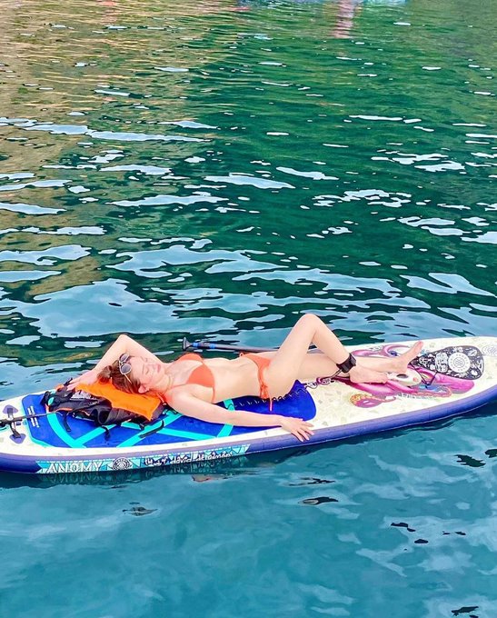 A woman in an orange bikini relaxing on a blue and white paddleboard floating on clear turquoise water.
