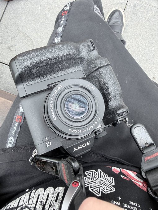 A first-person perspective photo showing a person wearing a black Sony Alpha mirrorless camera with a 16-50mm lens resting on their lap, which is covered in black clothing with red graphics.