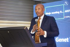 A man in a suit speaks into a microphone at a podium during an event, with text including "Multiple meetings," "AICI," "Genet," and "Sevoi" visible on screens behind him. The image relates to the post by illustrating a conference setting where topics like expanding market access across continents—particularly Africa, home to 17% of the global population—could be discussed in relation to IOTA's outreach.