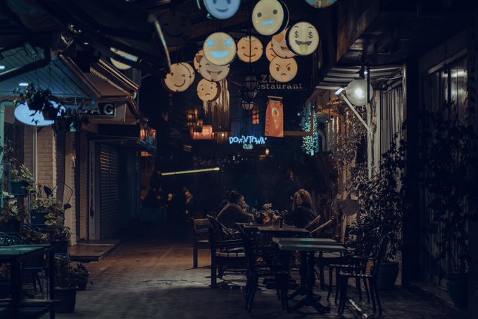 A dimly lit restaurant alleyway at night with hanging circular emoji-style lanterns, a 'DOWNTOWN' neon sign, and patrons seated at tables.