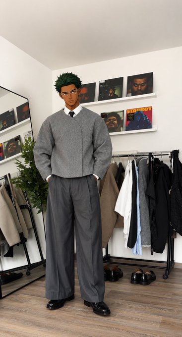 A person with green hair wearing a gray cardigan and wide-leg trousers poses in a room with clothing racks, vinyl records, and album covers including 'STAPBOY' on the shelves.