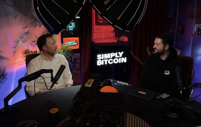 Two men sit across from each other at a table in the "Simply Bitcoin" studio, engaged in conversation while facing each other with professional microphones in front of them. The dimly lit space features a glowing "SIMPLY BITCOIN" sign in the background along with studio lighting and tropical decor, capturing an interview-style podcast moment featuring host and guest Mark Artymko discussing Ocean Mining.