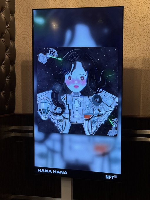 A digital screen displays a stylized cyberpunk NFT artwork of a girl with a helmet and robot companions, labeled 'HANA HANA' and 'NFT' at the bottom.