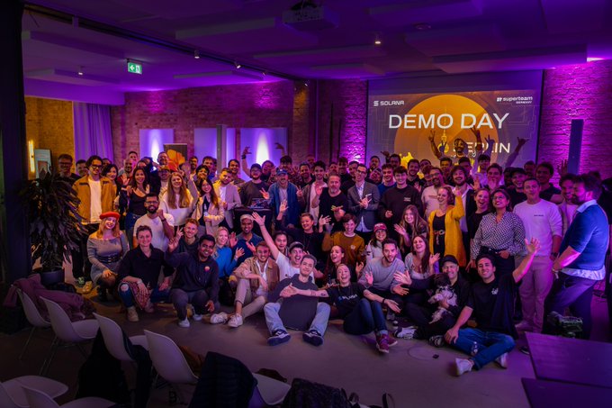 The image shows a large group of people gathered together for an event, possibly a Demo Day or similar gathering. The attendees are posing for a photo with their arms raised, creating a sense of unity and excitement among them. The room they are in appears to be a conference hall or a similar venue, as there is a projector screen visible in the background. The image also includes text that reads "Solana Summit Germany is landing in Berlin on June