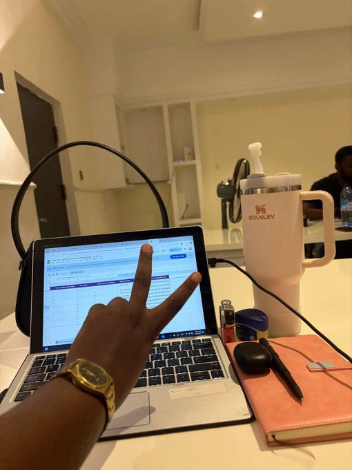 A person wearing a gold watch makes a peace sign in front of an open laptop displaying a spreadsheet, with a white Stanley tumbler, a lighter, AirPods case, a pen, and a pink notebook on the desk.