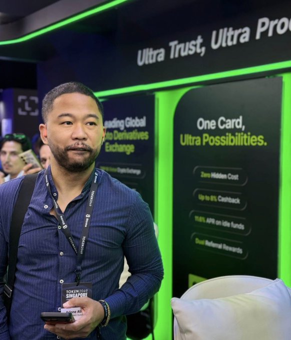 A man wearing a lanyard stands at the Token2049 Singapore event in front of a green-backed advertisement reading 'Ultra Trust, Ultra Pro' and 'One Card, Ultra Possibilities.'